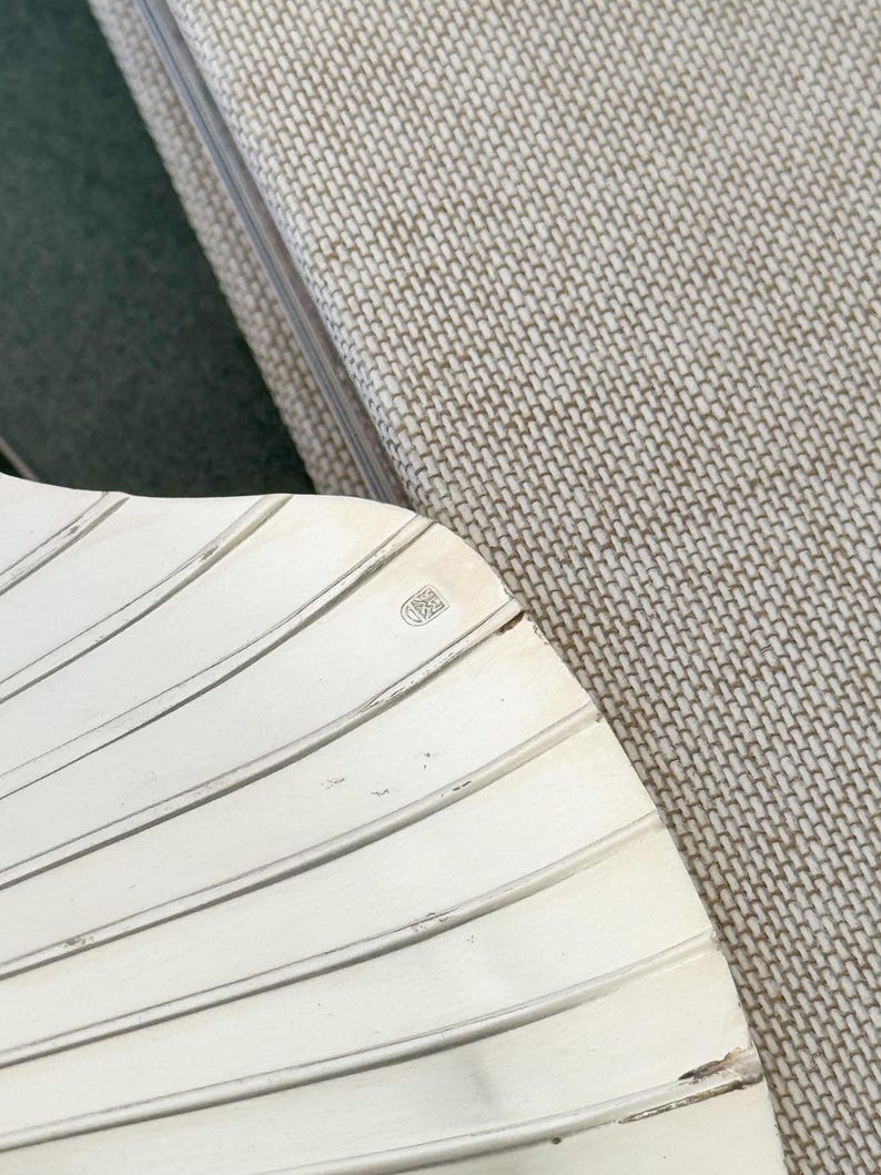 May include: A silver-colored, fan-shaped object with a ridged design. The object has a small emblem in the upper right corner. The background includes a textured, light-colored fabric and a dark green surface.