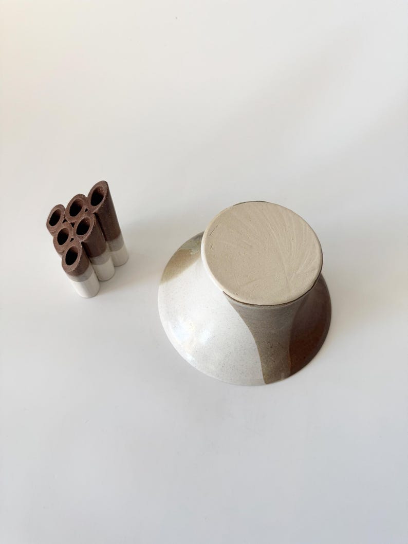 May include: A ceramic vase with a brown and white glaze. The vase has a wide, round base and a narrow neck. The vase is designed to hold multiple stems of flowers.