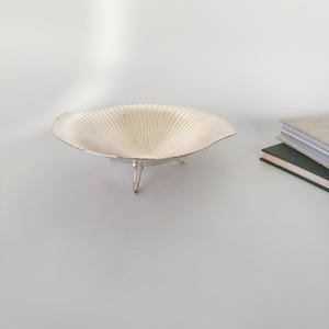May include: A silver-colored, shell-shaped bowl with a fluted design, resting on three small legs. The bowl has a wide, shallow form and is displayed on a light-colored surface. The background includes a stack of books.