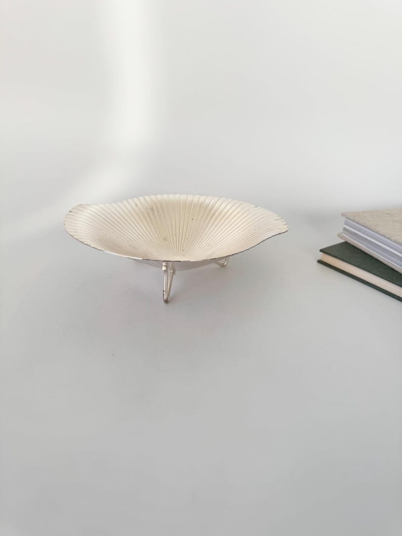 May include: A silver-colored, shell-shaped bowl with a fluted design, resting on three small legs. The bowl has a wide, shallow form and is displayed on a light-colored surface. The background includes a stack of books.