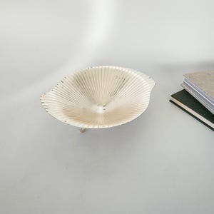 May include: A small, silver-colored decorative bowl with a unique, fan-like design. The bowl has a shallow, open shape and is supported by a delicate, curved base. The bowl is placed on a white surface, with a stack of books visible in the background.