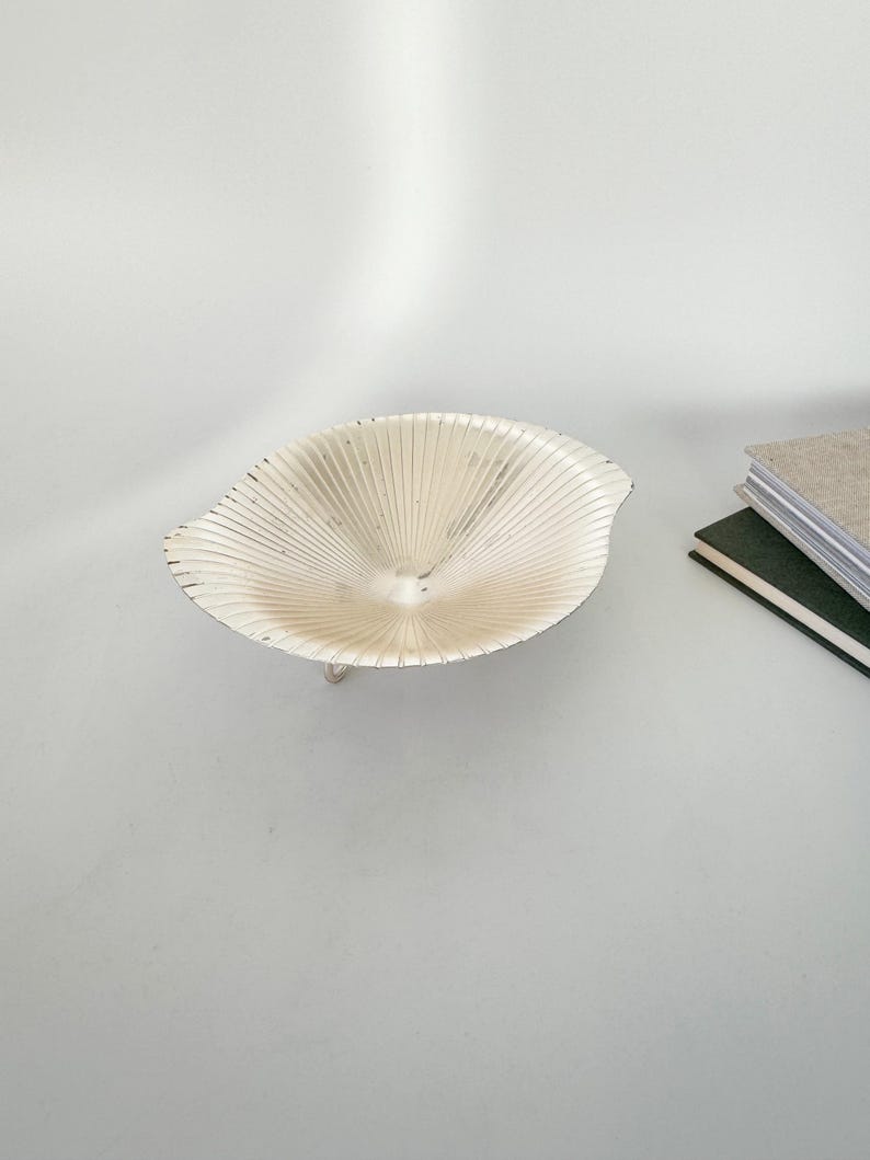May include: A small, silver-colored decorative bowl with a unique, fan-like design. The bowl has a shallow, open shape and is supported by a delicate, curved base. The bowl is placed on a white surface, with a stack of books visible in the background.