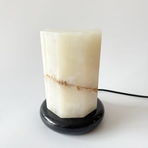May include: A white alabaster lamp with a black marble base. The lamp has a unique, geometric shape and features a natural, veined pattern.