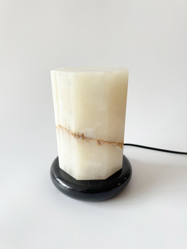 May include: A white alabaster lamp with a black marble base. The lamp has a unique, geometric shape and features a natural, veined pattern.
