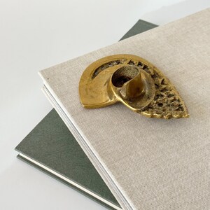 Op de afbeelding: Een vintage messing kandelaar in een schelpontwerp rust op een stapel boeken. De kandelaar heeft een getextureerd oppervlak en een centrale beker voor de kaars. De boeken hebben een lichtbeige en donkergroene kaft.