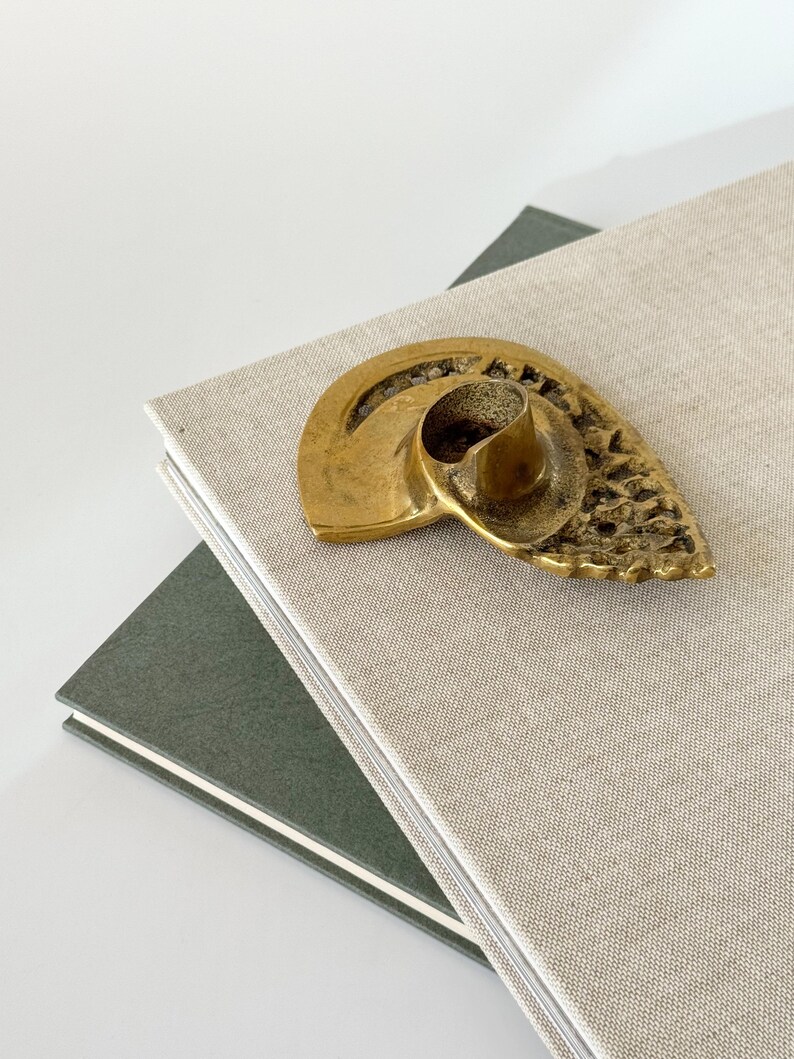 Op de afbeelding: Een vintage messing kandelaar in een schelpontwerp rust op een stapel boeken. De kandelaar heeft een getextureerd oppervlak en een centrale beker voor de kaars. De boeken hebben een lichtbeige en donkergroene kaft.