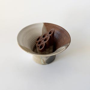 May include: A brown and white ceramic vase with a unique design. The vase has a wide, flared opening and a narrow base. It features a brown, textured, circular insert in the center.