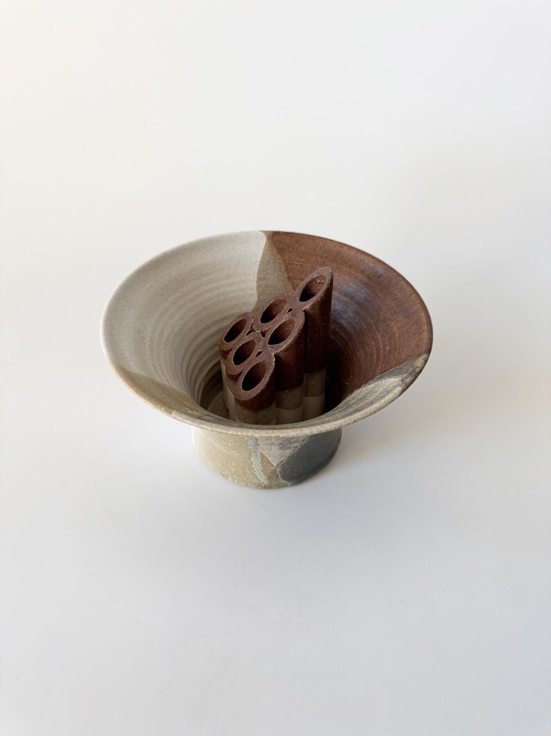 May include: A brown and white ceramic vase with a unique design. The vase has a wide, flared opening and a narrow base. It features a brown, textured, circular insert in the center.
