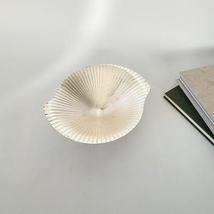 May include: A silver-colored, fan-shaped decorative bowl with a textured surface. The bowl has a central indentation and is positioned on a white surface. Two books are visible in the background.