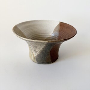 May include: A small, ceramic bowl with a flared rim and a brown, gray, and white glaze. The bowl has a unique, hand-crafted look.