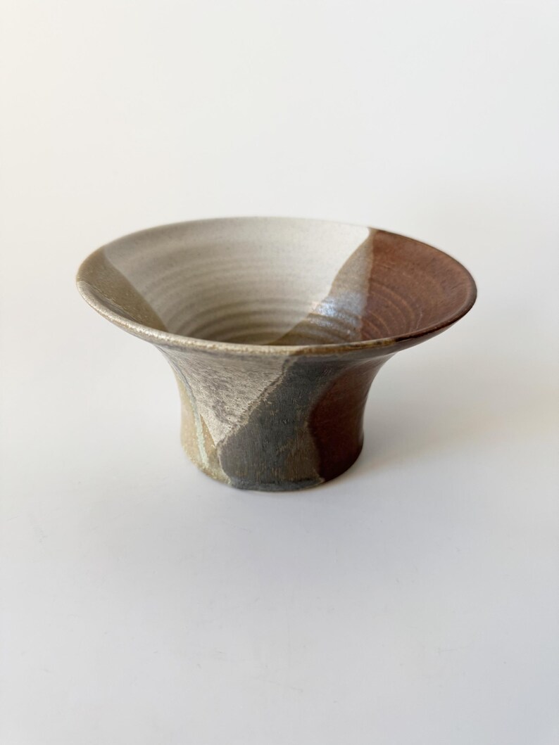 May include: A small, ceramic bowl with a flared rim and a brown, gray, and white glaze. The bowl has a unique, hand-crafted look.