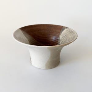 May include: A small, white ceramic vase with brown and gray glaze. The vase has a flared opening and a rounded base.