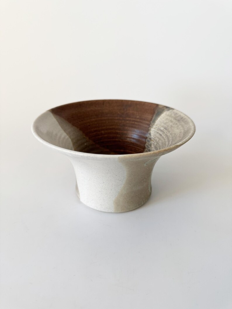 May include: A small, white ceramic vase with brown and gray glaze. The vase has a flared opening and a rounded base.