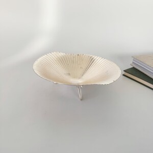 May include: A decorative, silver-colored bowl with a fan-like design. The bowl is supported by three small, silver legs. The bowl is empty and sits on a light gray surface. A stack of books is visible in the background.