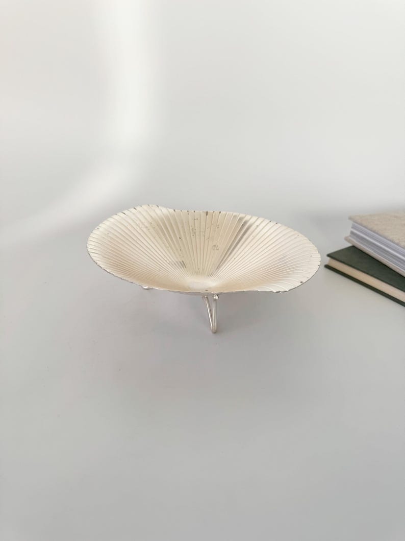 May include: A decorative, silver-colored bowl with a fan-like design. The bowl is supported by three small, silver legs. The bowl is empty and sits on a light gray surface. A stack of books is visible in the background.