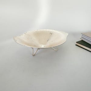 May include: A decorative, silver-colored bowl with a unique, fan-like design. The bowl is supported by three slender, silver legs. The bowl's interior has a textured, ridged pattern. The bowl is likely used for decorative purposes or to hold small items.