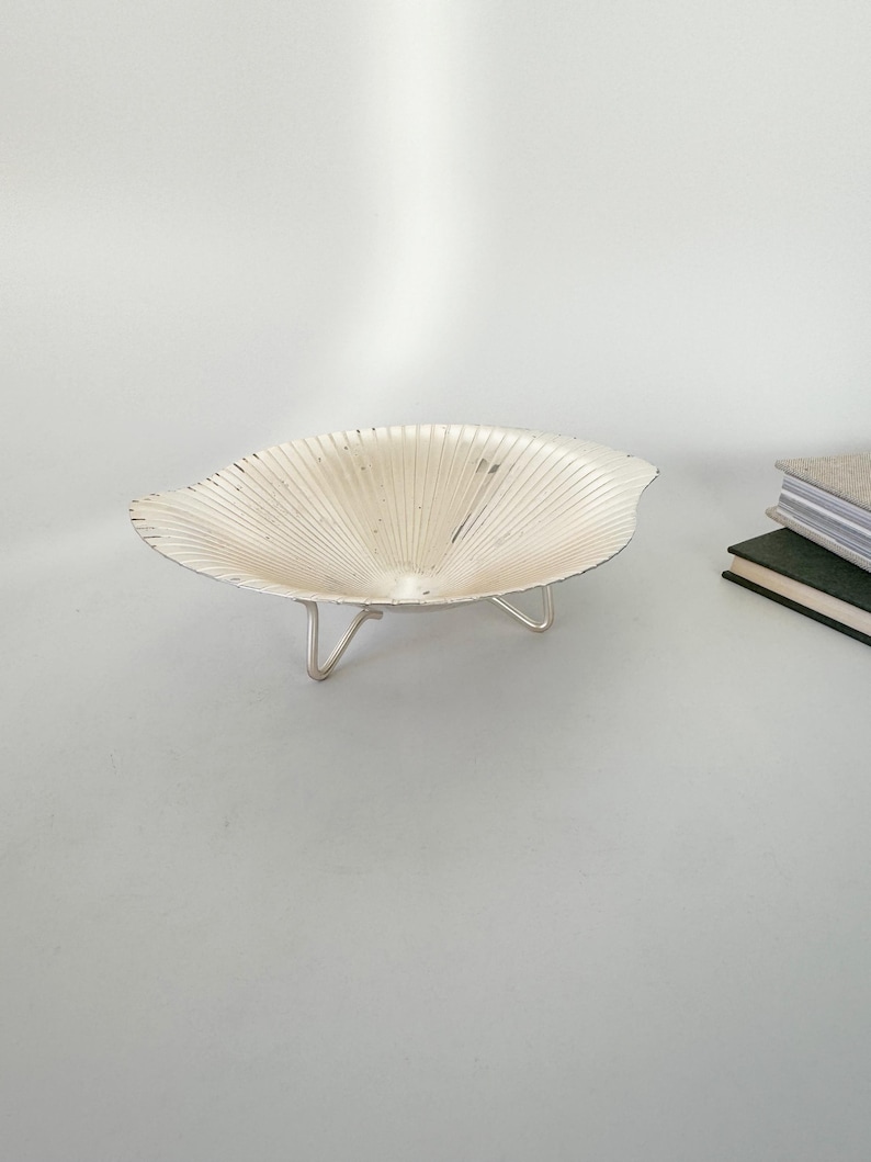 May include: A decorative, silver-colored bowl with a unique, fan-like design. The bowl is supported by three slender, silver legs. The bowl's interior has a textured, ridged pattern. The bowl is likely used for decorative purposes or to hold small items.