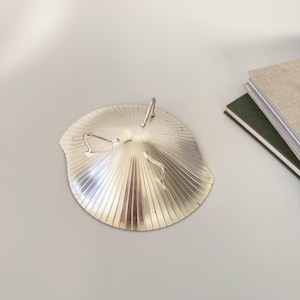 May include: A silver-colored, fan-shaped dish with a ridged design. The dish has three curved metal accents and a central, upright metal piece. The dish is on a light gray surface.