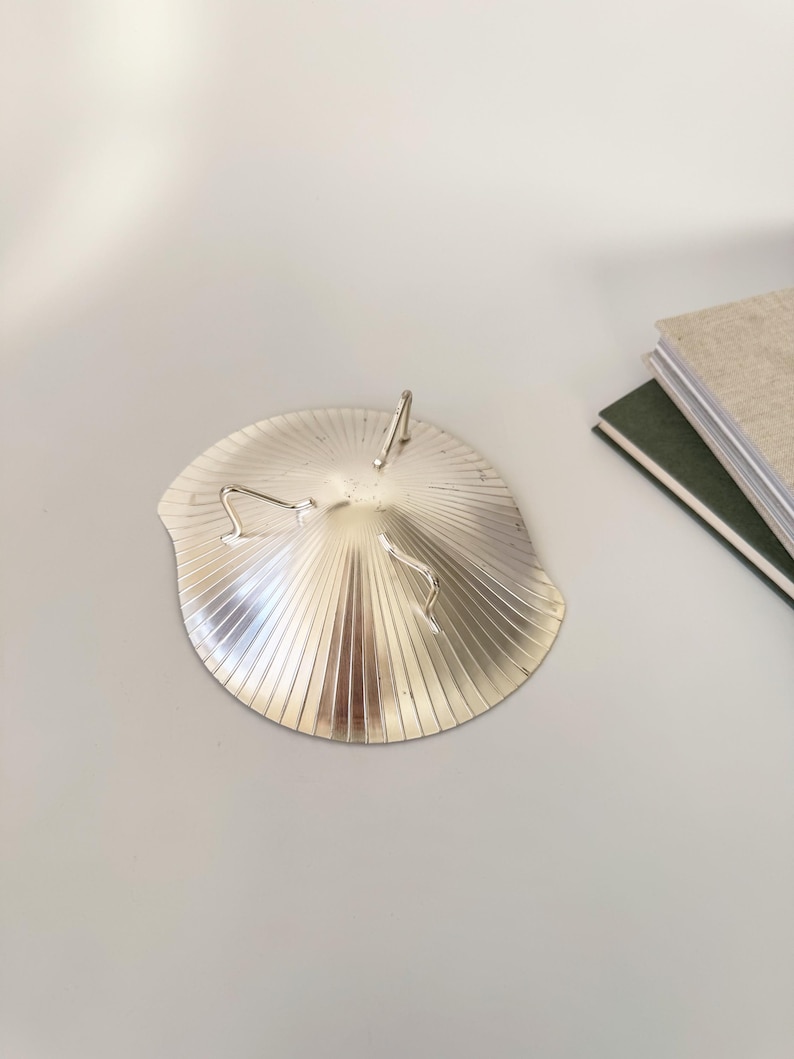 May include: A silver-colored, fan-shaped dish with a ridged design. The dish has three curved metal accents and a central, upright metal piece. The dish is on a light gray surface.