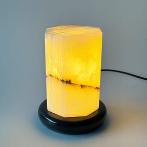 May include: A white alabaster table lamp with a black base. The lamp is lit and the light shines through the alabaster, creating a warm glow.