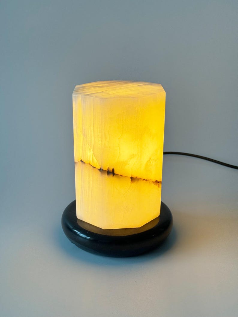 May include: A white alabaster table lamp with a black base. The lamp is lit and the light shines through the alabaster, creating a warm glow.