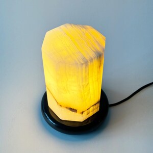 May include: A white alabaster lamp with a geometric shape and a black base. The lamp is lit and casting a warm glow.