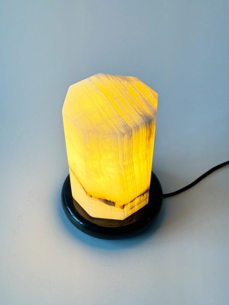 May include: A white alabaster lamp with a geometric shape and a black base. The lamp is lit and casting a warm glow.