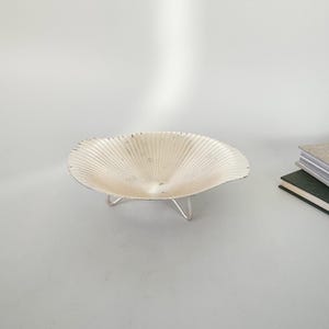 May include: A cream-colored, shell-shaped bowl with a textured surface, supported by three silver-colored legs. The bowl has a fluted design radiating from the center. The background is a plain white surface.
