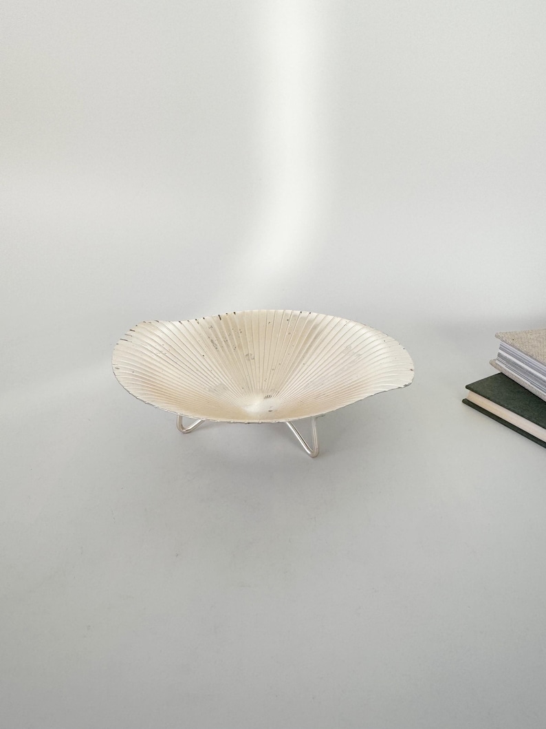 May include: A cream-colored, shell-shaped bowl with a textured surface, supported by three silver-colored legs. The bowl has a fluted design radiating from the center. The background is a plain white surface.