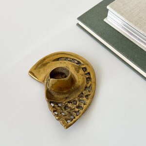 Op de afbeelding: Een vintage messing kandelaar in de vorm van een zeeschelp. De houder heeft een centrale beker voor de kaars en ingewikkelde opengewerkte details langs de randen. Twee boeken staan op de achtergrond.