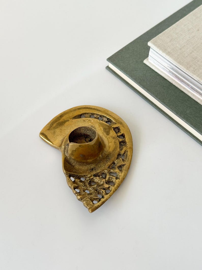 Op de afbeelding: Een vintage messing kandelaar in de vorm van een zeeschelp. De houder heeft een centrale beker voor de kaars en ingewikkelde opengewerkte details langs de randen. Twee boeken staan op de achtergrond.