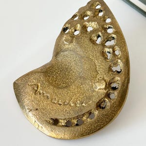 Op de afbeelding: Een goudkleurige, abstracte sculptuur met een getextureerd oppervlak en onregelmatige gaten. Het stuk heeft een gebogen, organische vorm, mogelijk een decoratief object of kunstwerk. De sculptuur heeft een vintage of antieke uitstraling.