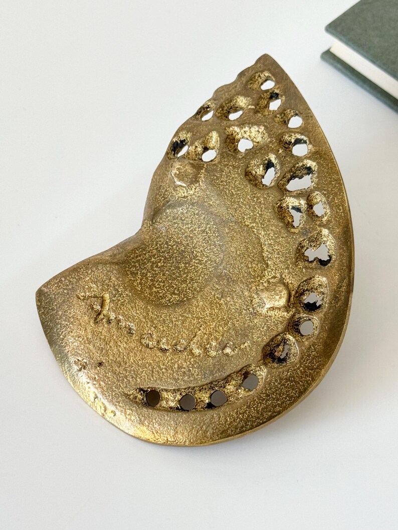 Op de afbeelding: Een goudkleurige, abstracte sculptuur met een getextureerd oppervlak en onregelmatige gaten. Het stuk heeft een gebogen, organische vorm, mogelijk een decoratief object of kunstwerk. De sculptuur heeft een vintage of antieke uitstraling.