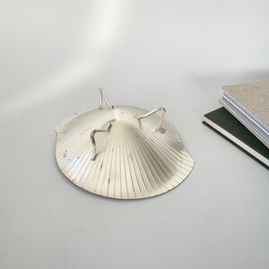 May include: A silver-colored, shell-shaped dish with three curved, silver legs. The dish has a ridged, fan-like pattern radiating from the center. A stack of books is visible in the background. This decorative dish is suitable for serving or display.