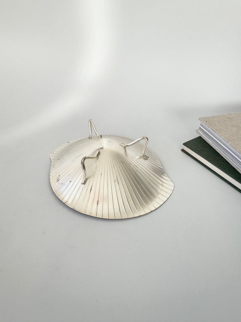 May include: A silver-colored, shell-shaped dish with three curved, silver legs. The dish has a ridged, fan-like pattern radiating from the center. A stack of books is visible in the background. This decorative dish is suitable for serving or display.