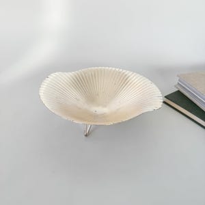 May include: A cream-colored, shell-shaped bowl with a ribbed design and three silver legs. The bowl is made of metal and has a slightly distressed finish. The bowl is a decorative item.