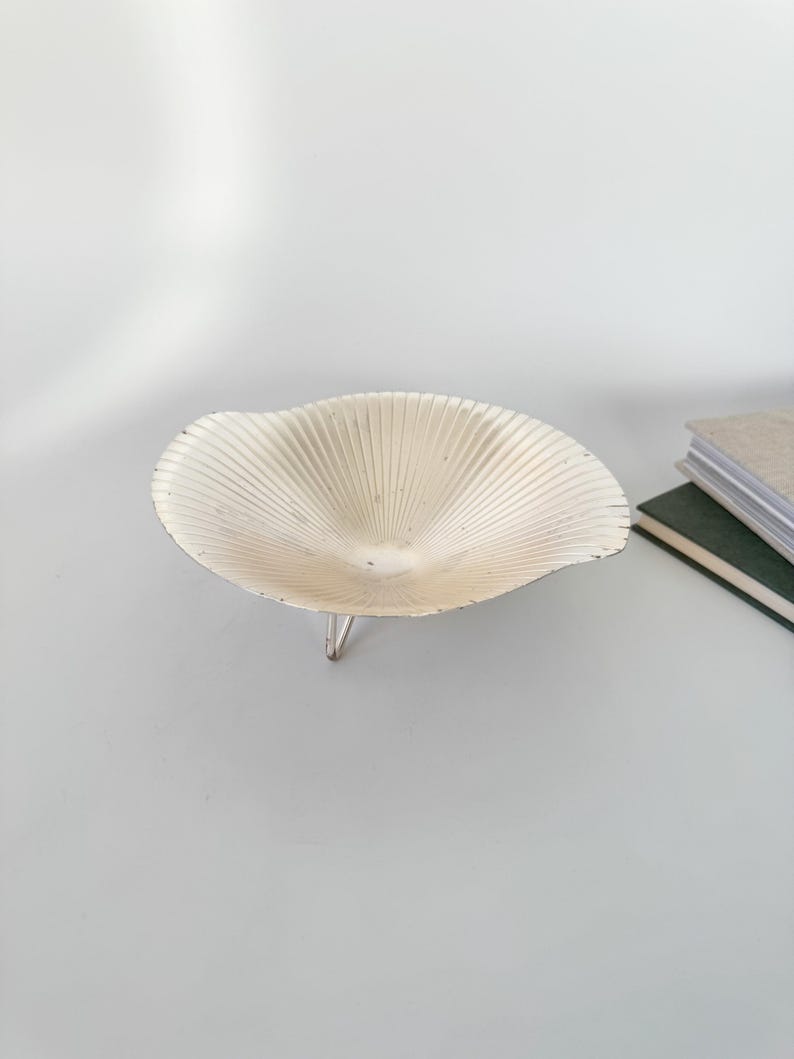 May include: A cream-colored, shell-shaped bowl with a ribbed design and three silver legs. The bowl is made of metal and has a slightly distressed finish. The bowl is a decorative item.