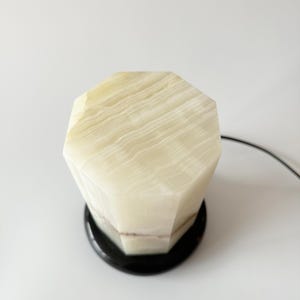 May include: A white onyx table lamp with a geometric, faceted base and a black base. The lamp is on a white surface.