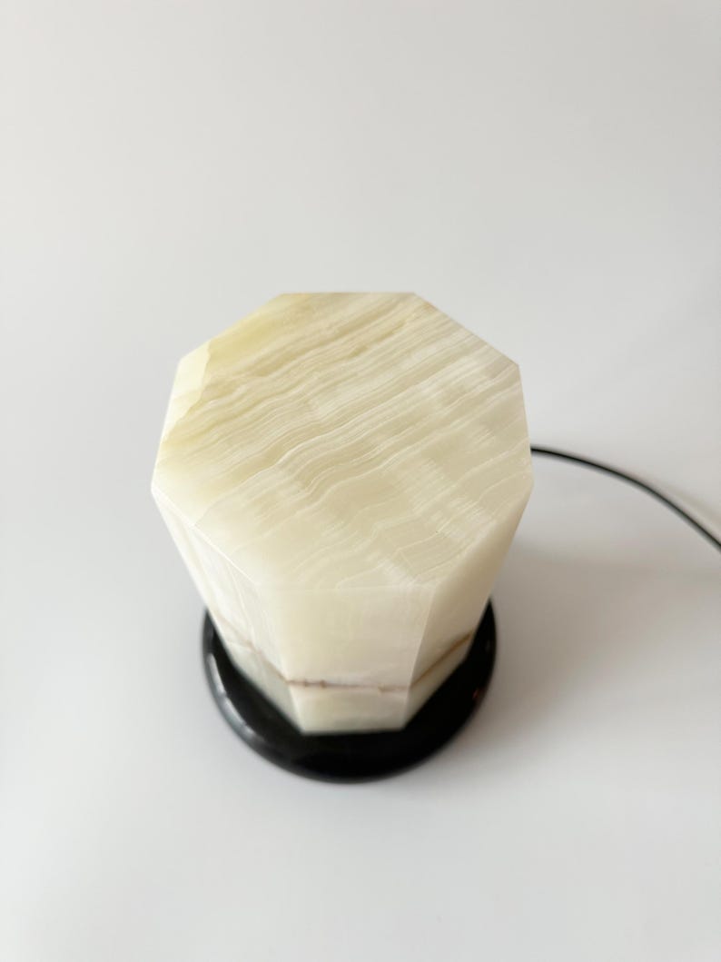 May include: A white onyx table lamp with a geometric, faceted base and a black base. The lamp is on a white surface.