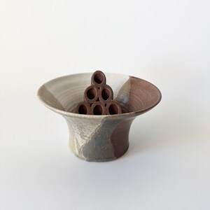 May include: A brown and white ceramic bowl with a geometric design. The bowl has a wide, flared rim and a narrow base. A brown ceramic flower frog is inside the bowl.