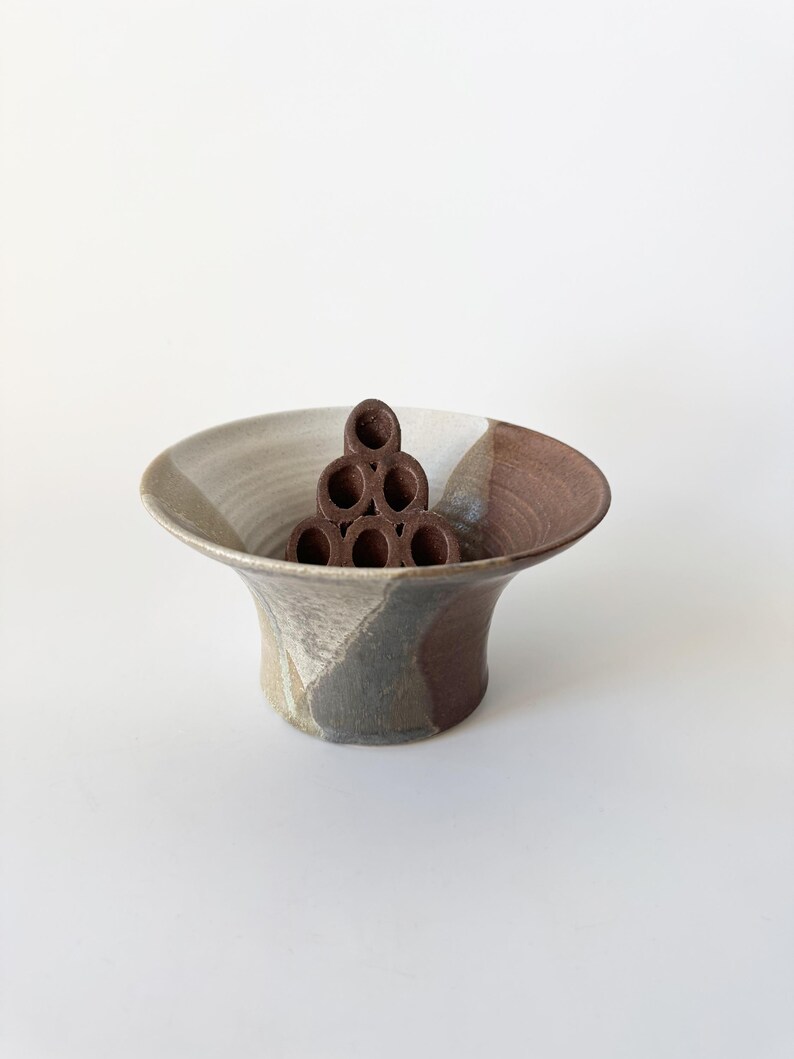 May include: A brown and white ceramic bowl with a geometric design. The bowl has a wide, flared rim and a narrow base. A brown ceramic flower frog is inside the bowl.