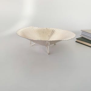 May include: A silver-colored, decorative bowl with a scalloped design and three small legs. The bowl has a shallow, open shape and a textured surface. The bowl is likely used for decorative purposes or to hold small items.