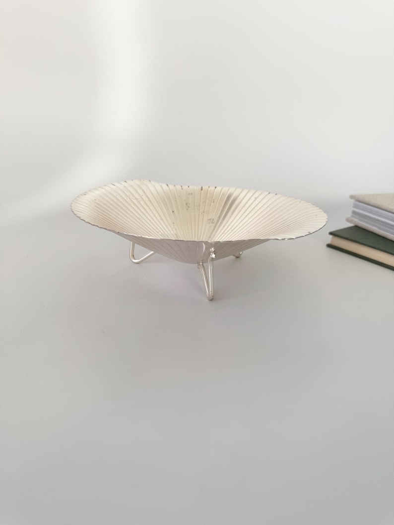 May include: A silver-colored, decorative bowl with a scalloped design and three small legs. The bowl has a shallow, open shape and a textured surface. The bowl is likely used for decorative purposes or to hold small items.