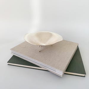 May include: A decorative, off-white seashell-shaped bowl with three silver legs, resting on top of two hardback books. The top book is beige, and the bottom book is dark green. The background is a solid white.
