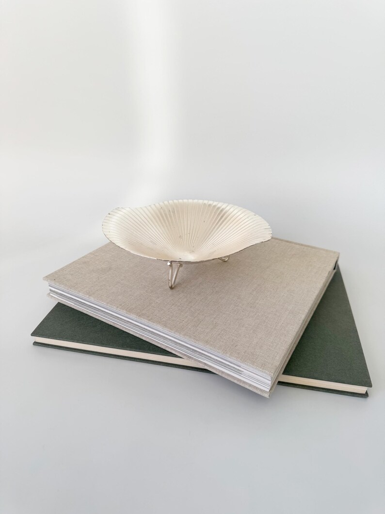 May include: A decorative, off-white seashell-shaped bowl with three silver legs, resting on top of two hardback books. The top book is beige, and the bottom book is dark green. The background is a solid white.