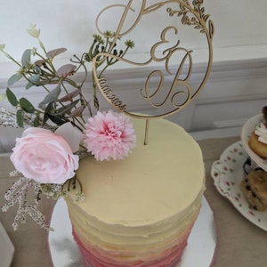 Könnte beinhalten: Eine weiße und rosafarbene Ombre-Hochzeitstorte mit einem Holz-Tortenaufsatz mit den Initialen "E & B" und dem Datum "10.05.2023". Die Torte ist mit rosa Blumen verziert.