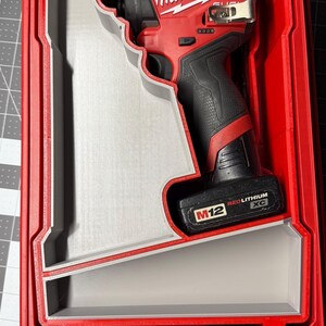 以下が含まれることがあります： カスタムフィットの赤とグレーのケースに入った、赤いMilwaukee M12インパクトドライバー。インパクトドライバーは赤と黒で、Milwaukeeのロゴが見えます。ケースは、グレーの内装と赤いトリムで、ツールを安全に保持するように設計されています。