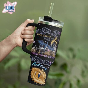 May include: A black stainless steel tumbler with a black and white graphic of a dragon and a castle. The tumbler has a black handle and a straw. The text "Riders Quadrant" and "I Will Not Die Today" is printed on the tumbler. The name "Andarna" is printed on the bottom of the tumbler.