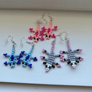 May include: Three pairs of beaded dangle earrings featuring animal shapes. One pair is light blue and dark blue lizards, one pair is pink lizards, and one pair is gray, black, and white opossums. Each earring has a silver-toned metal hook.