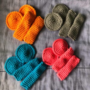 Puede incluir: Conjunto de artículos de ganchillo hechos a mano en varios colores: naranja, turquesa, coral y verde oliva. El conjunto presenta posavasos circulares y fundas rectangulares, todos dispuestos sobre una superficie de tela gris. Ideal para la decoración del hogar.